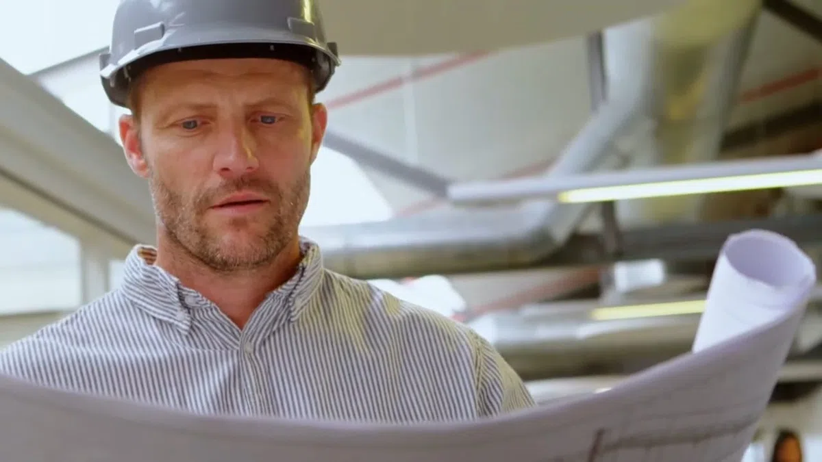 Licensed plumber in a hard hat reviewing job plans near overhead piping for Should You Repipe Your Goodyear Home Before Selling? The Buyer Inspection Reality